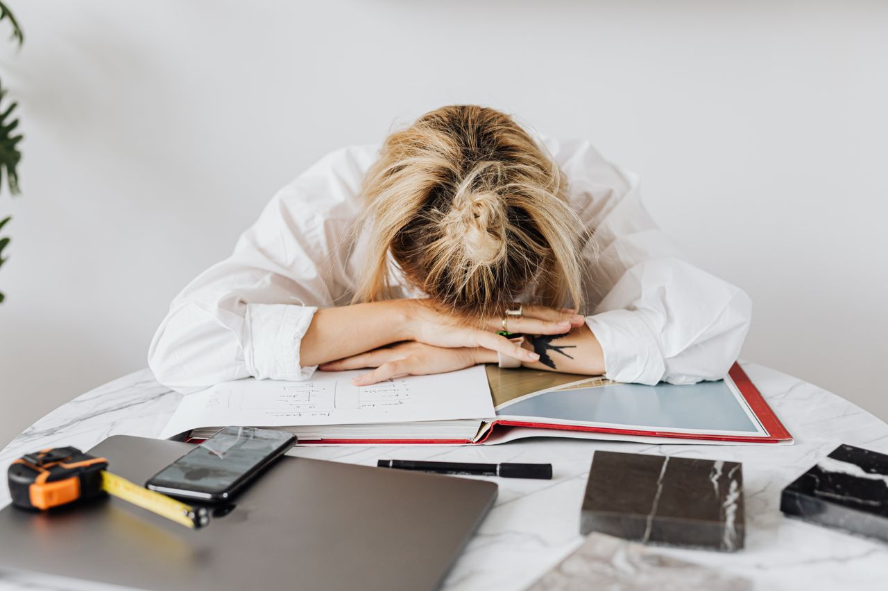 Tired woman in white shirt resting her head on a desk filled with work materials.